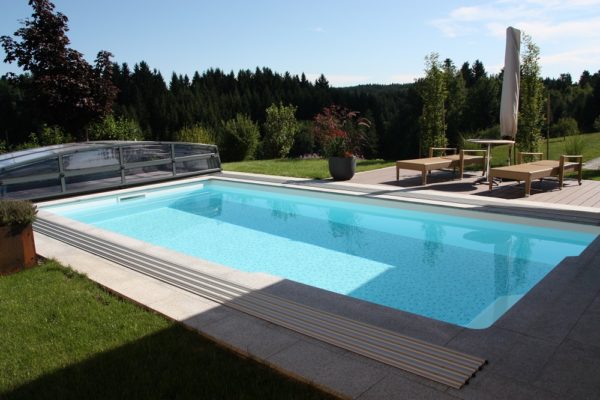 Das Rechteck-Schwimmbecken Ozean vereint zeitlosen Stil mit langlebiger Schönheit. Das Bild zeigt ein Schwimmbecken Ozean mit geöffneten Cabriodom. Im Hintergrund ist ein Wald und Sräucher zu sehen. Am Beckenrand steht ein kleiner Tisch mit zwei Liegestühle und ein geschlossener Schirm.