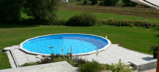 Cranpool Royal besticht durch seine stabile Konstruktion. Das Bild zeigt ein rundes Schwimmbecken Royal das komplett in die Erde eingebaut ist. Im Vordergrund des Beckens wurden graue Steinplatten verlegt. Im Hintergrund ist eine Wiese mit anschließenden Acken.