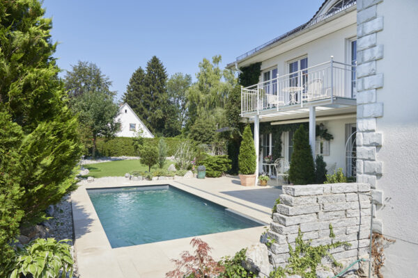 Pool im Garten, das rechteckige Schwimmbecken von Cranpool ist eingegraben und somit auf einer Ebene mit der Terasse . Die Terasse wurde so verlängert, dass rund ums Pool ein Steg gebaut wurde. Im hintergreund erkennt man einen Garten von einem Einfamilien Haus